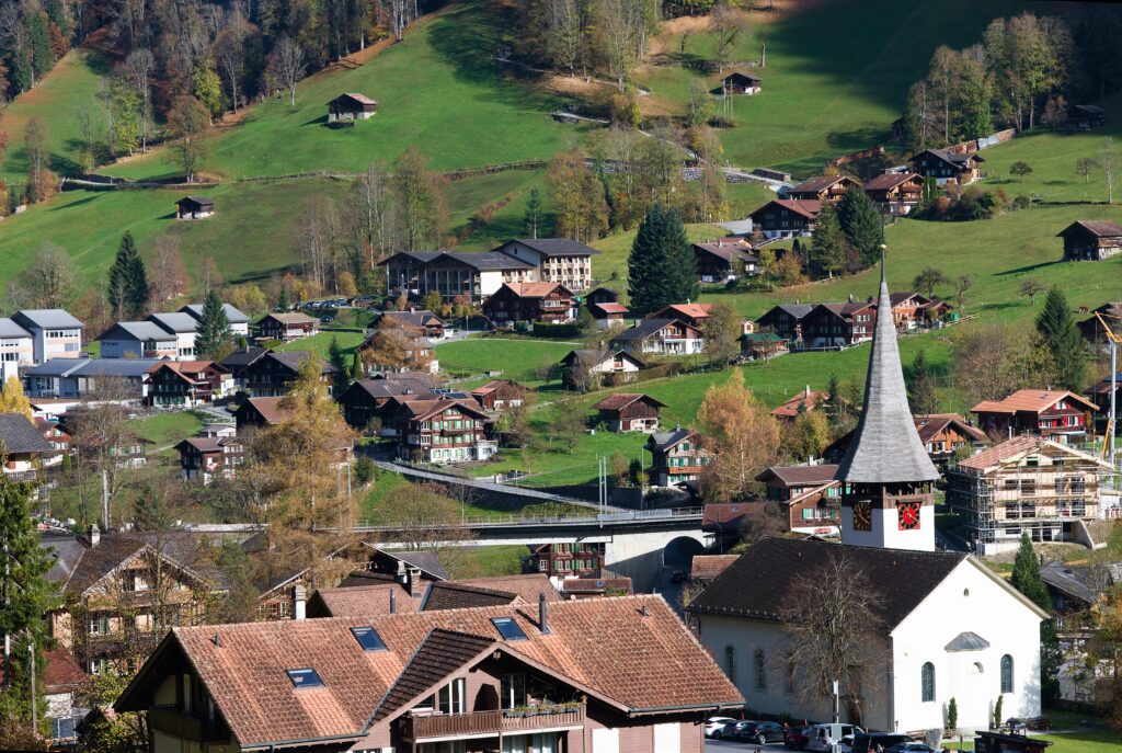 Charming Swiss village scene with church, traditional houses, and lush green hills in autumn sunlight.